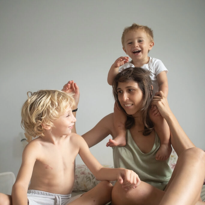 Fotografía de familia en Barcelona: Madre feliz jugando con sus dos hijos, un bebé en hombros y un niño, capturando momentos de alegría en casa.